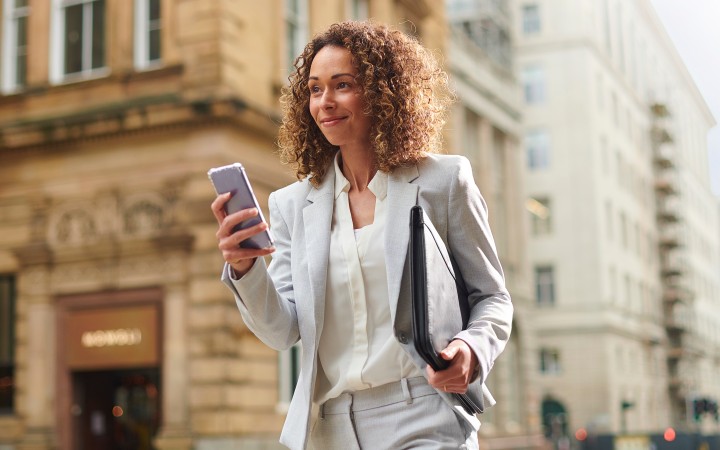 A woman walks down a street with a briefcase under her arm and a smartphone in her hand.
