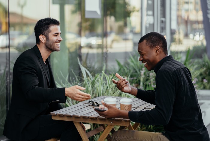 Two people are sitting at a table outside having a coffee and laughing together.