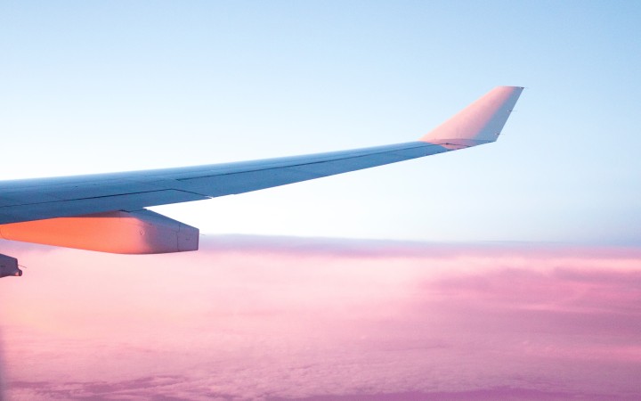 The wing of an airplane is seen above the clouds, flying into a sunrise.